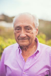 Warm portrait of an elderly man in a natural setting wearing a lavender shirt.